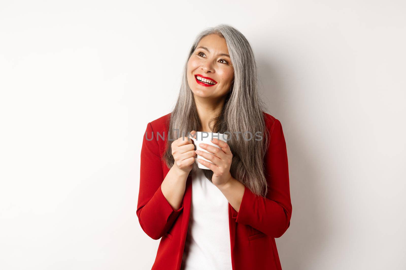 Business people concept. Smiling asian businesswoman having coffee break, holding warm mug and looking upper left corner dreamy, white background.