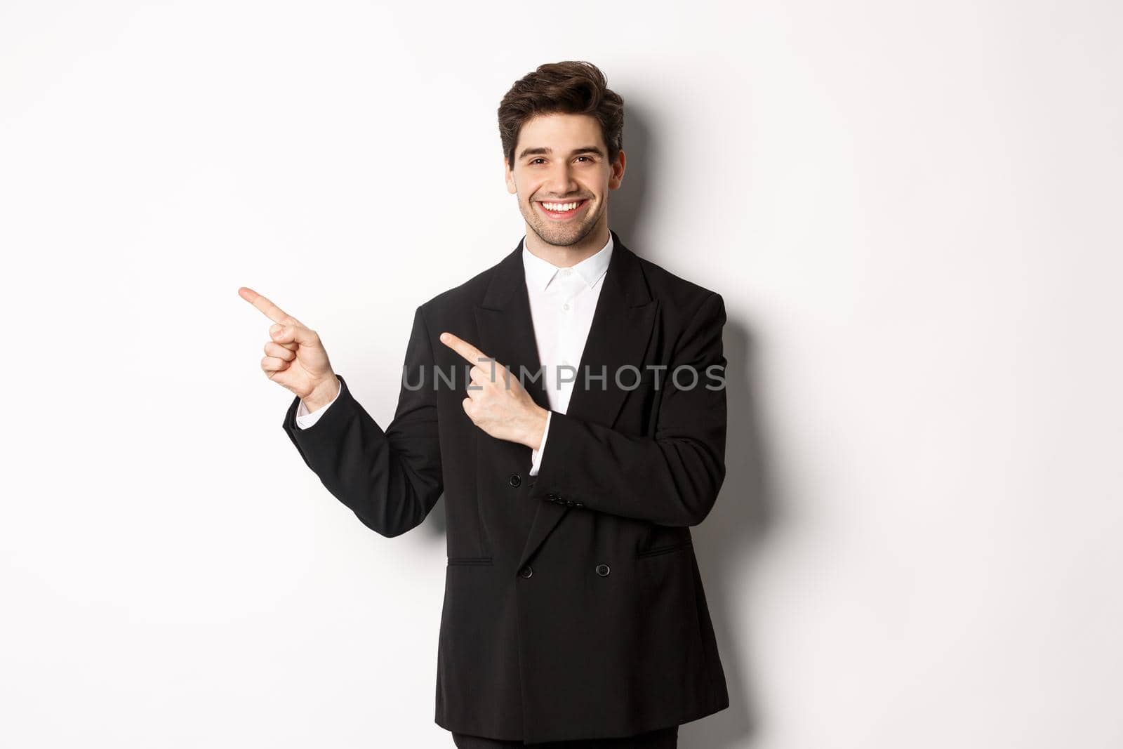 Portrait of successful, handsome businessman in suit, pointing fingers at upper left corner and smiling, showing advertisement, standing over white background.