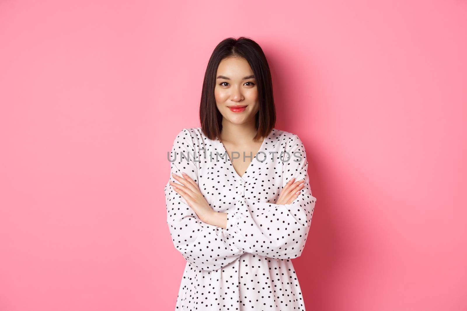 Beautiful asian female model standing in dress, cross arms on chest and smiling at camera, standing over pink background.