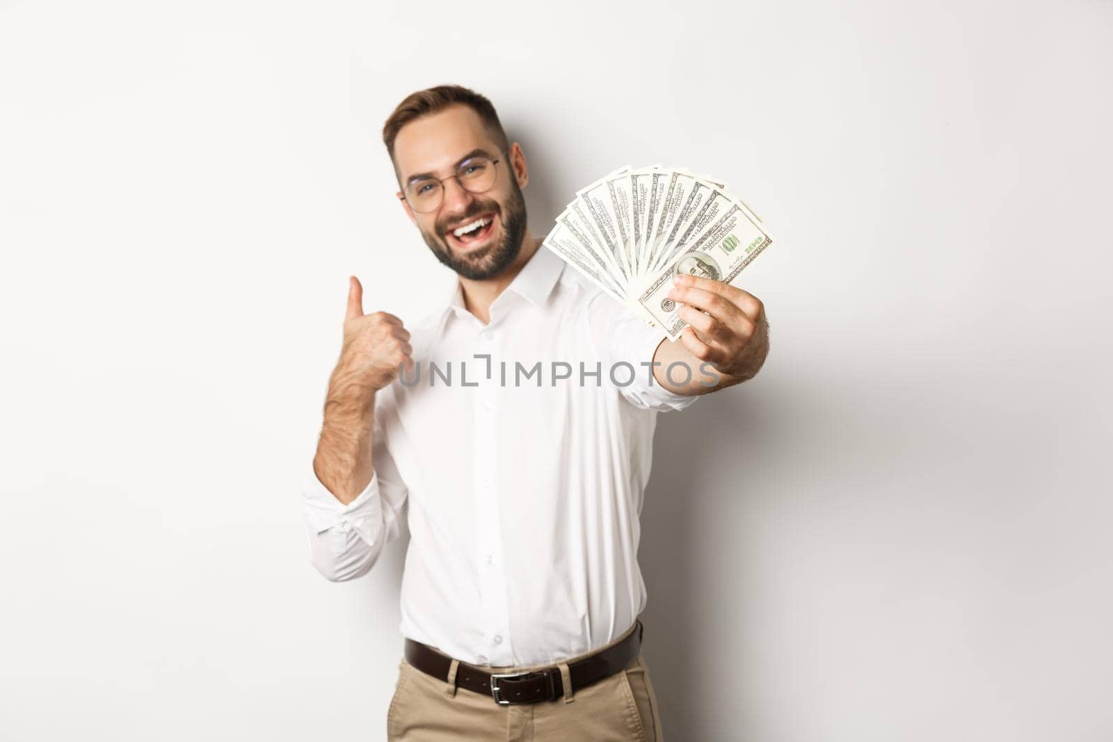 Successful businessman showing money dollars and thumbs-up, smiling satisfied, standing over white background by Benzoix