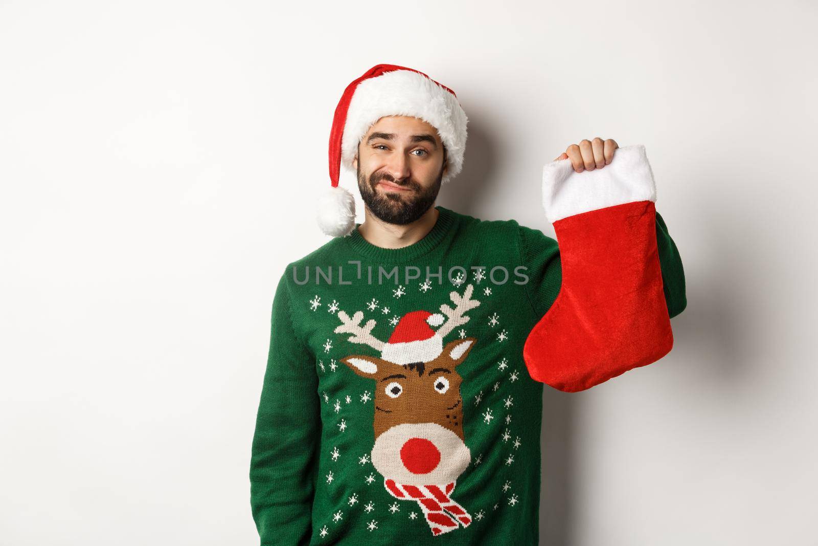 Xmas and winter holidays concept. Displeased guy disappointed in gift in Christmas sock, standing upset in Santa hat against white background.