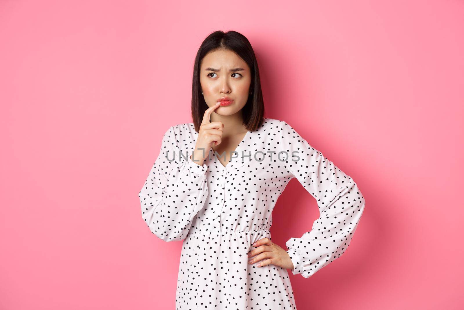 Troubled asian girl making decision, frowning and touching lip while thinking, looking unsure at upper left corner and choosing, standing over pink background.