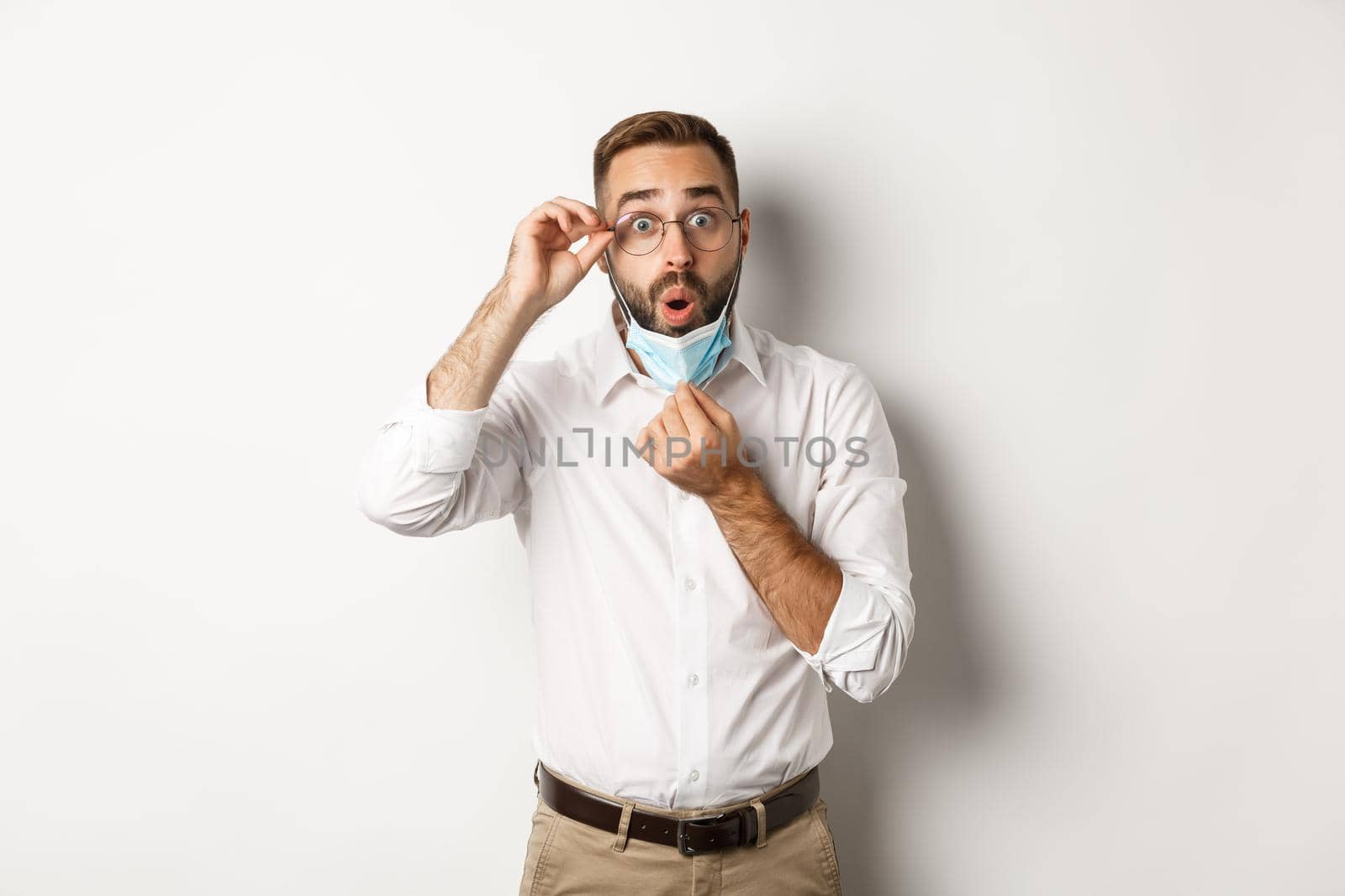 Covid-19, social distancing and quarantine concept. Impressed business man take off medical mask, looking surprised, standing over white background.