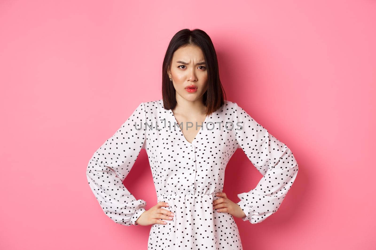 Angry asian woman staring at camera frustrated, holding hands on waist and waiting for explanations, standing in dress against pink background by Benzoix