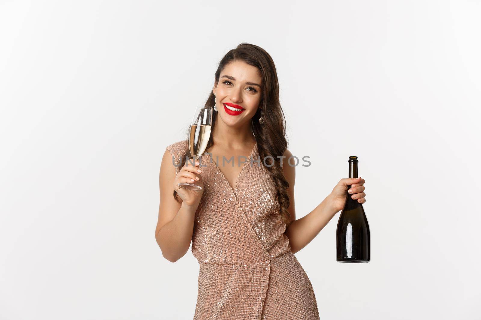 Winter holidays celebration concept. Image of happy young woman in luxury dress, drinking champagne from glass, holding bottle, New Year party, white background.