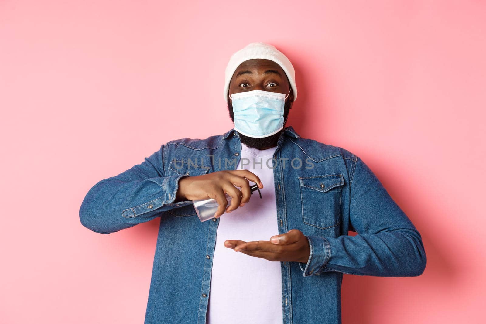 Covid-19, lifestyle and lockdown concept. Smiling african-american man in face mask cleaning hands with sanitizer, using antiseptic and looking at camera, pink background.