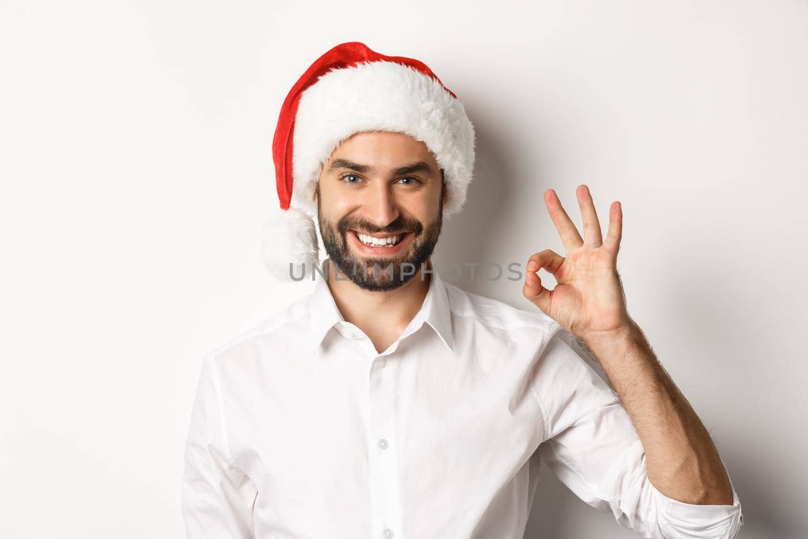 Party, winter holidays and celebration concept. Confident man in santa hat showing okay sign, approve and like, white background by Benzoix