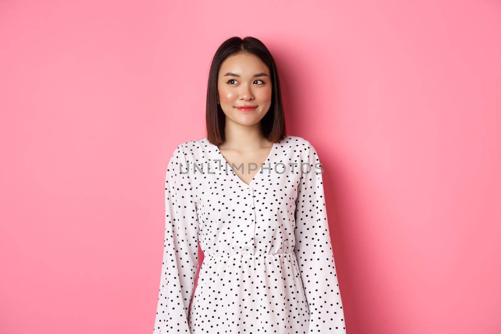 Cute asian woman smiling, looking left at copy space, standing on romantic pink background.