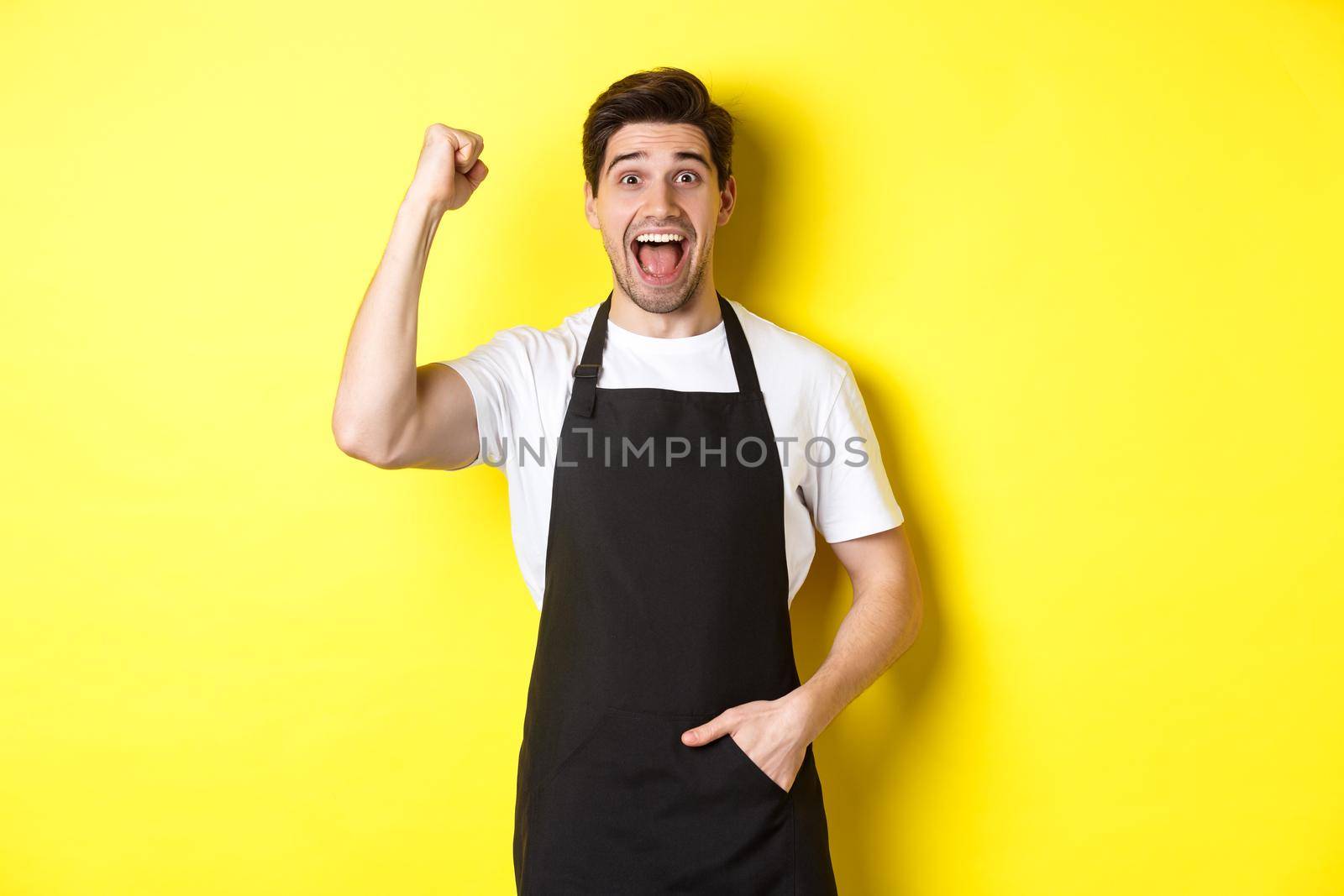 Cheerful seller making fist pump, rejoicing and triumphing, standing in black apron against yellow background by Benzoix