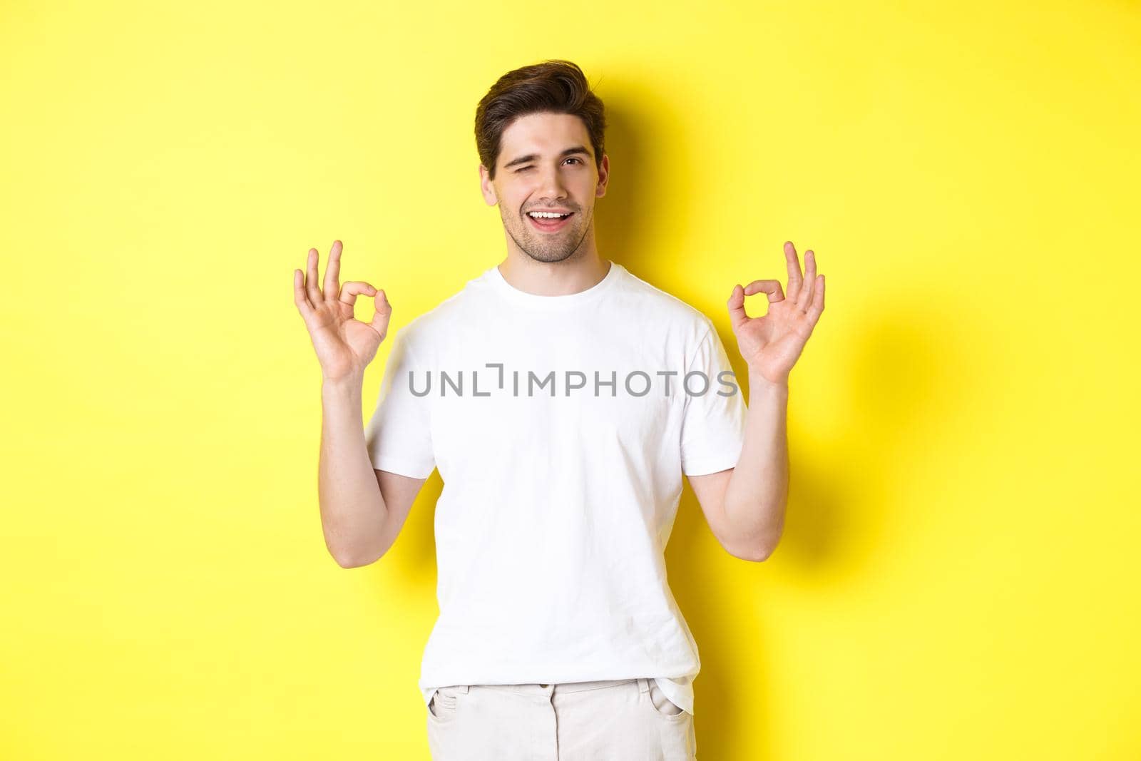 Confident handsome man winking, showing okay signs in approval, like something good, standing over yellow background.