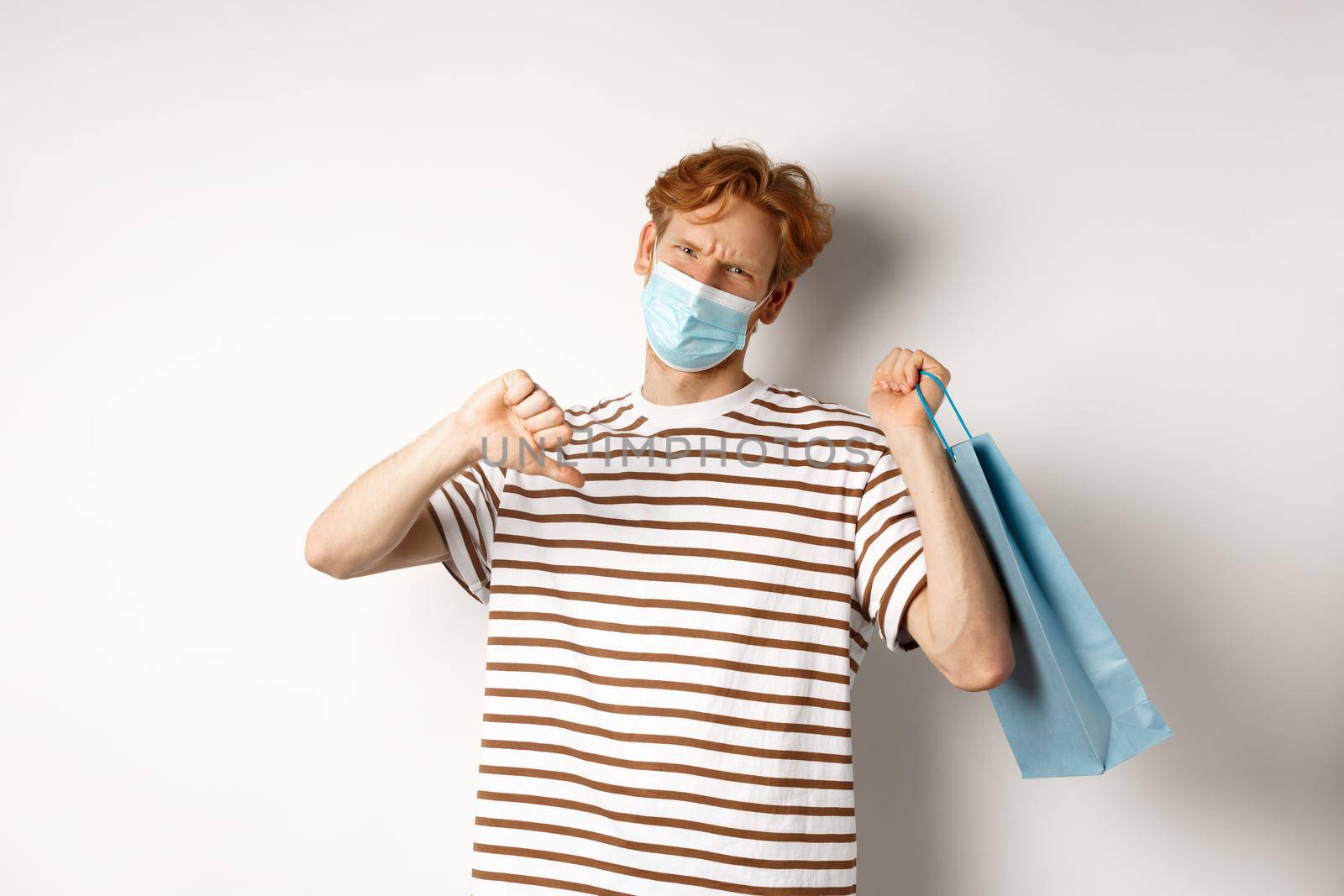 Concept of social distancing and shopping. Disappointed young man with red hair, wearing face mask, holding shopping bag and showing thumb-down from dislike, disapprove store by Benzoix