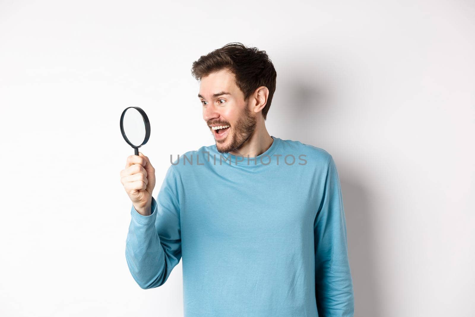Handsome smiling man looking through magnifying glass with amazed face, found interesting promotion, standing on white background.