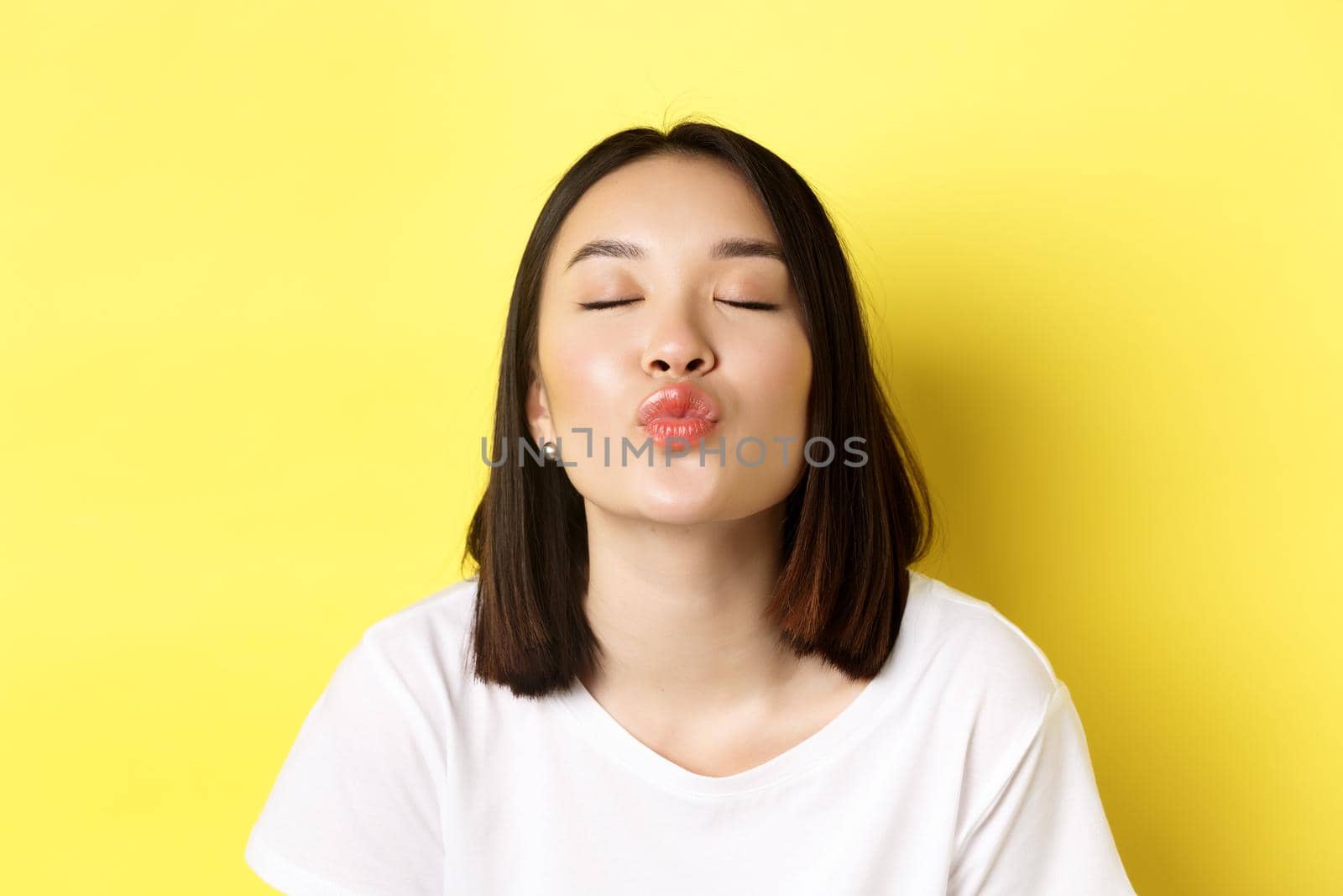 Valentines day concept. Close up of cute asian girl pucker lips and close eyes for kiss, standing romantic against yellow background.