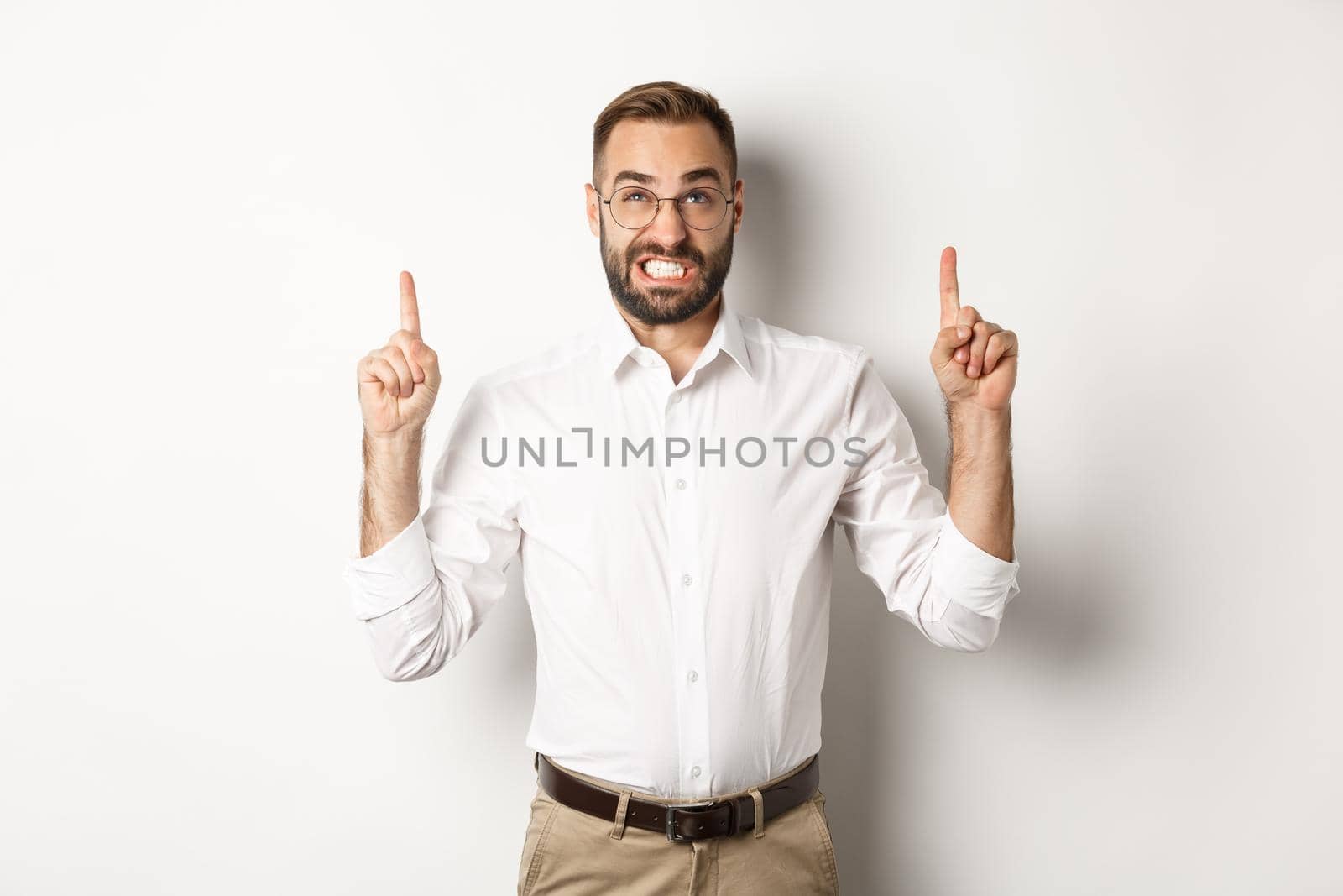 Doubtful and worried man looking and pointing fingers up, grimacing hesitant, standing over white background.