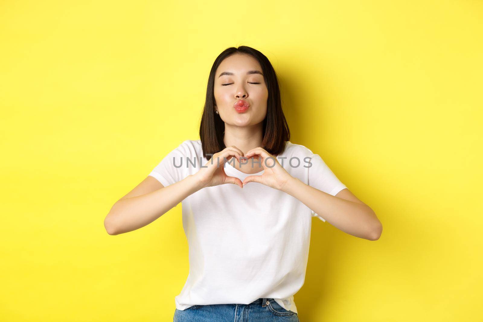 Beautiful asian woman showing I love you heart gesture, smiling at camera, standing against yellow background. Concept of valentines day and romance by Benzoix