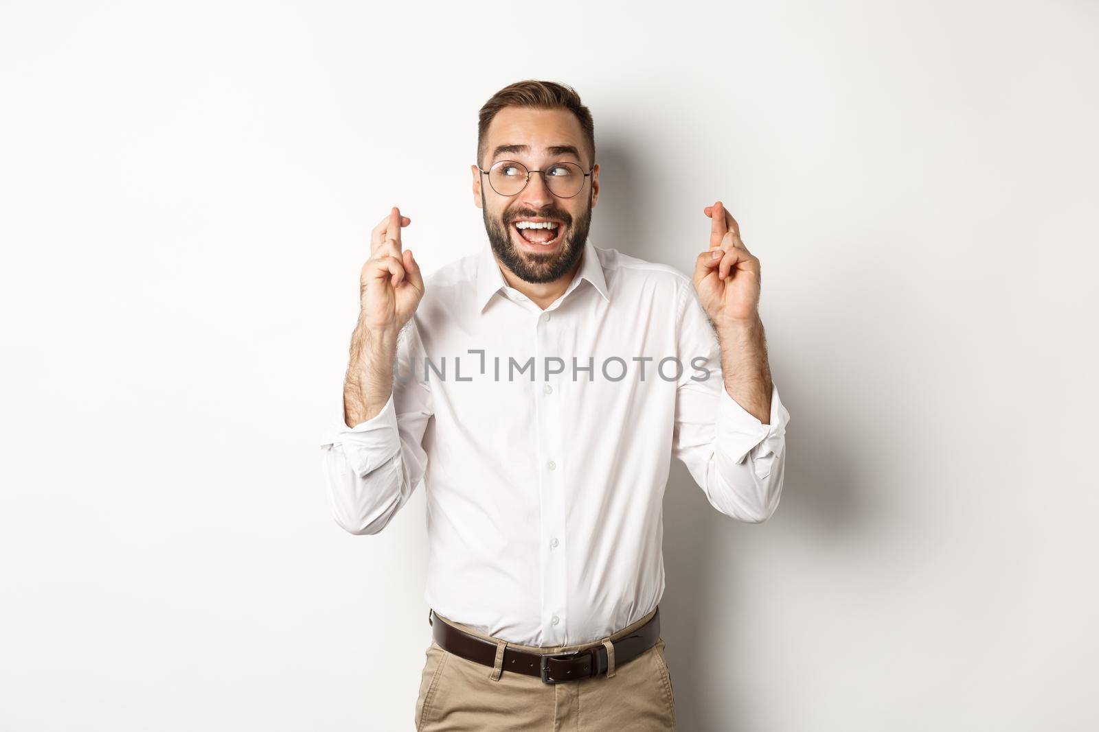 Excited and hopeful businessman making a wish, cross fingers and waiting, standing over white background.