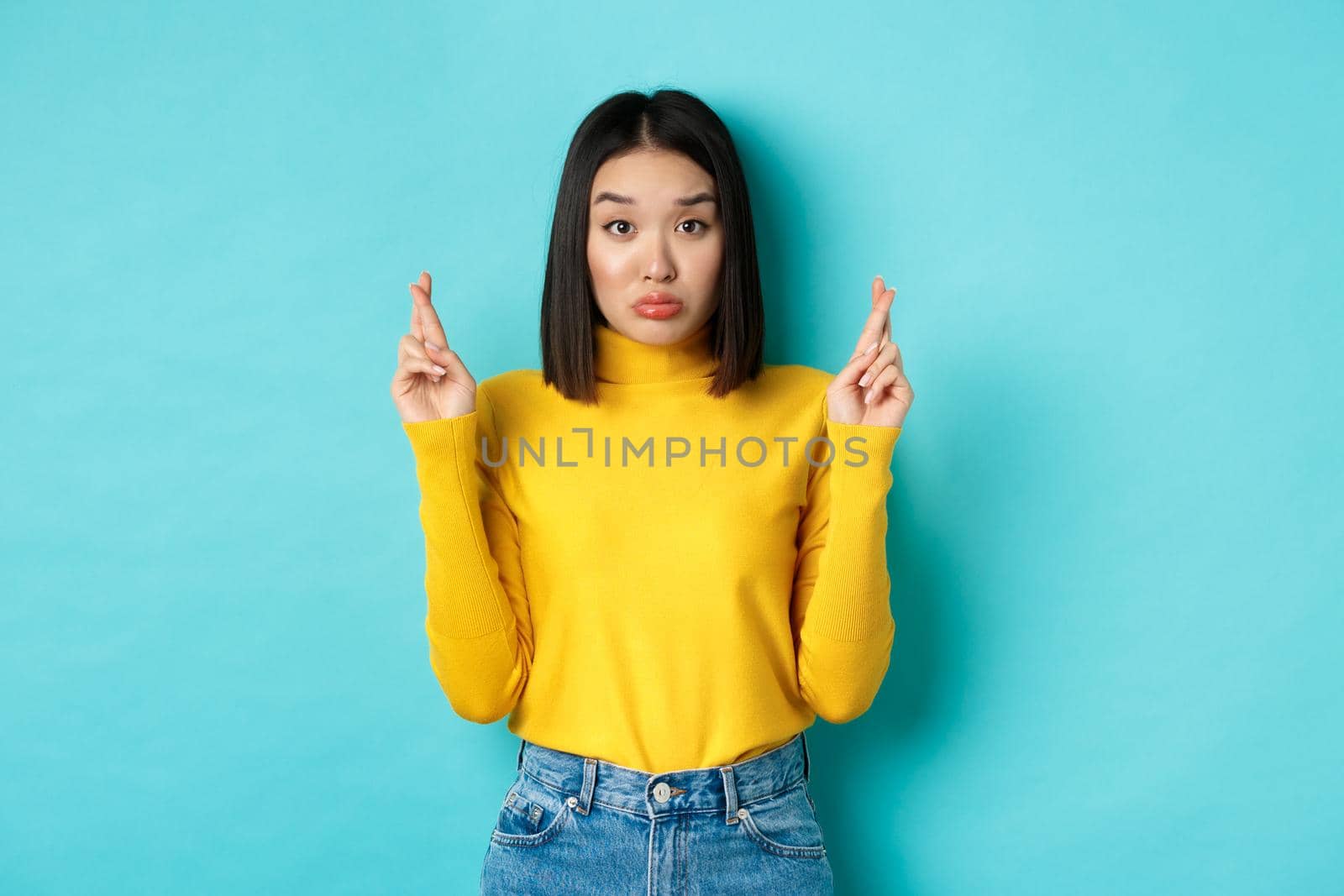 Silly hopeful asian girl making wish, pucker lips and looking at camera with dreamy glance, cross fingers for good luck, standing over blue background.