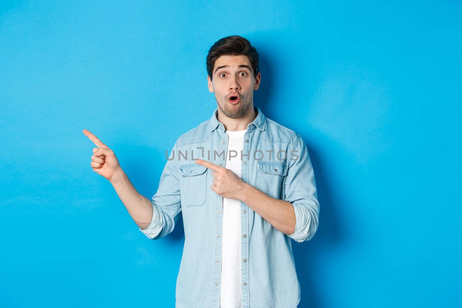 Portrait of surprised adult man in casual clothes showing announcement, pointing fingers left and looking amazed, standing against blue background.