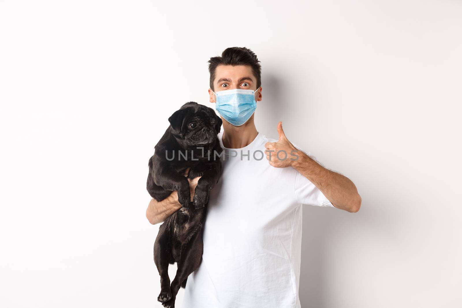 Covid-19, animals and quarantine concept. Young man in medical mask holding cute black pug dog, showing thumb up, like and approve, standing over white background.