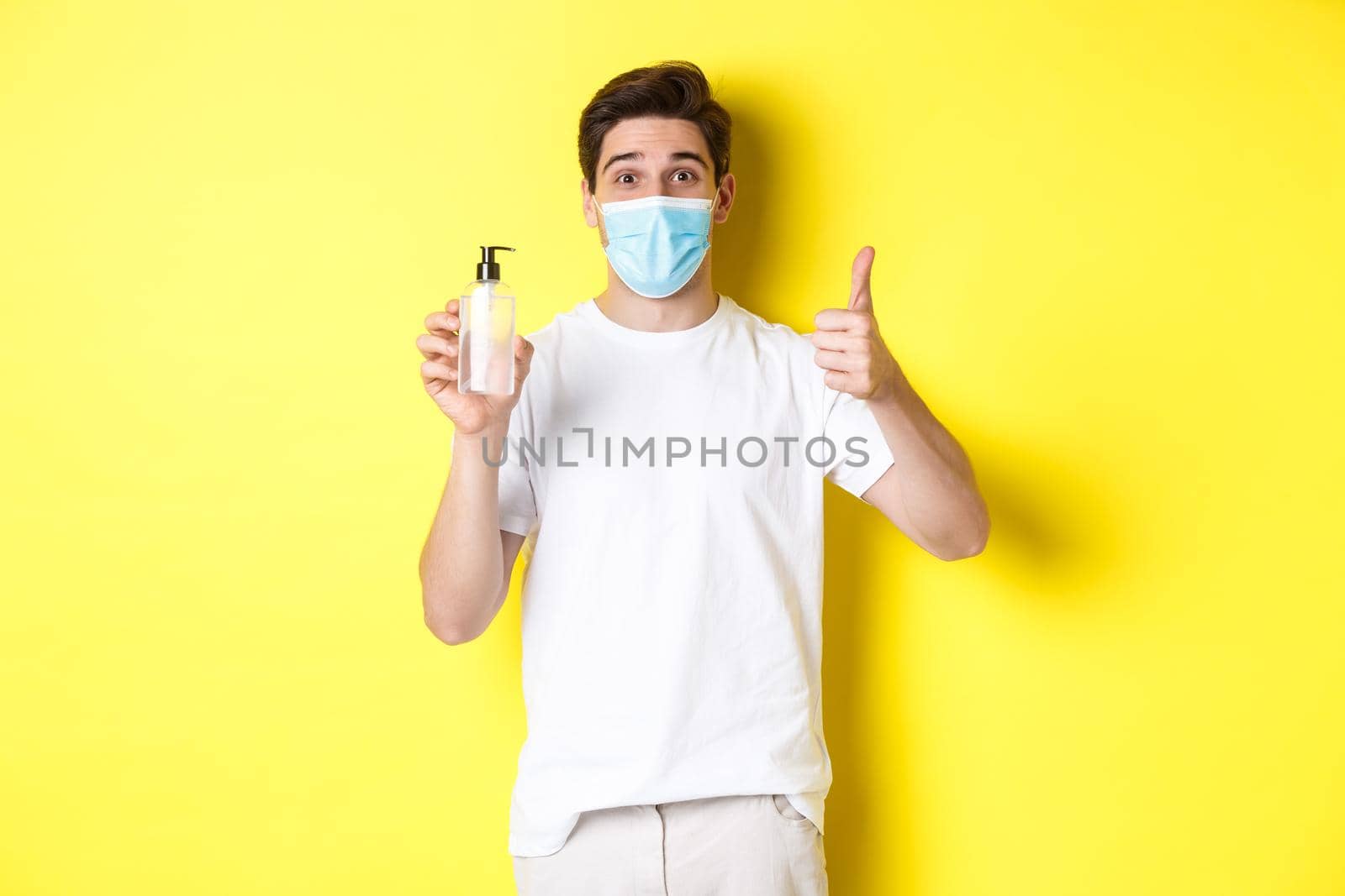 Concept of covid-19, quarantine and lifestyle. Satisfied young man in medical mask showing good hand sanitizer, thumbs up and recommending antiseptic, yellow background by Benzoix