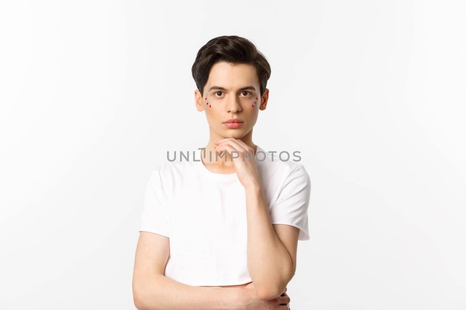 Image of attractive gay man in white t-shirt, having glitter on face and looking at camera serious, standing over white background.