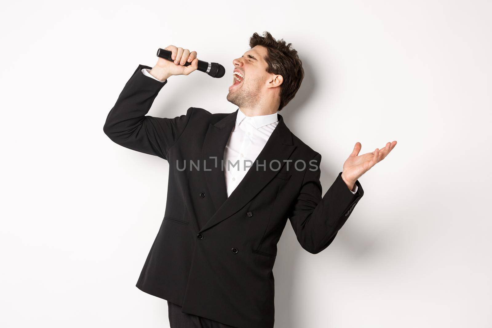 Portrait of handsome man singing a song with passion, standing in black suit, holding microphone and performning, posing over white background by Benzoix