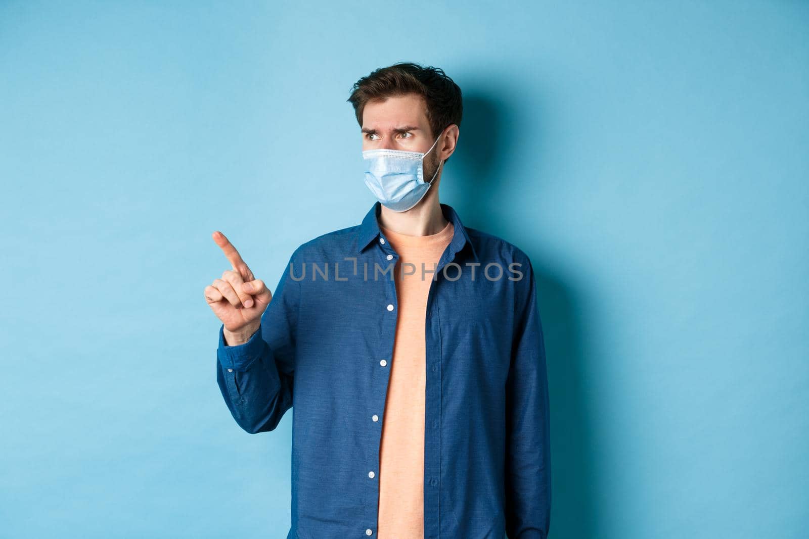 Covid-19 and healthcare concept. Confused and doubtful man in face mask frowning, pointing and looking at empty space, standing on blue background by Benzoix