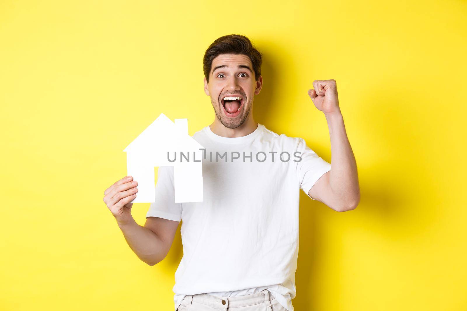 Real estate concept. Cheerful man showing paper house model and making fist pump, paid mortgage, yellow background by Benzoix