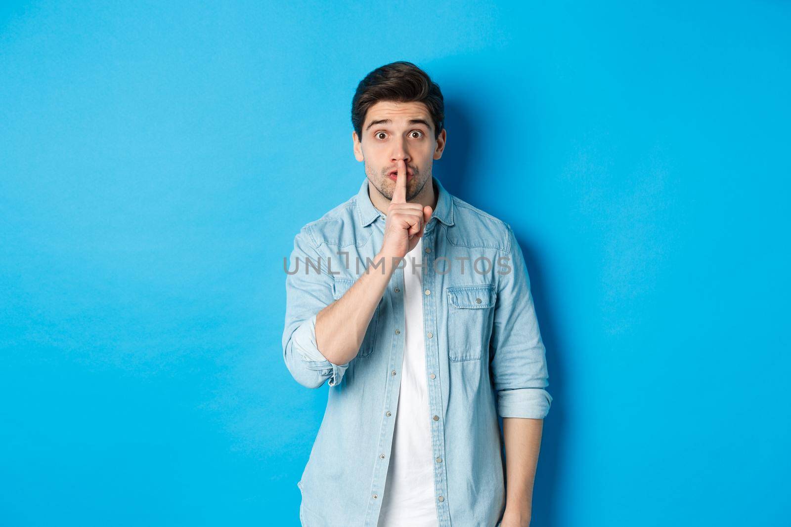 Portrait of excited man asking to keep quiet, showing hush taboo sign and looking nervously at camera, standing against blue background by Benzoix