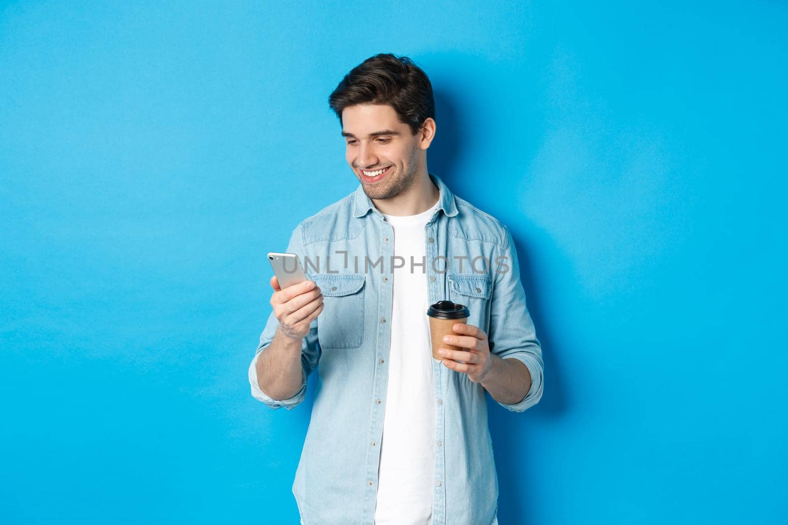 Handsome modern guy reading message on cell phone and drinking coffee, standing against blue background.