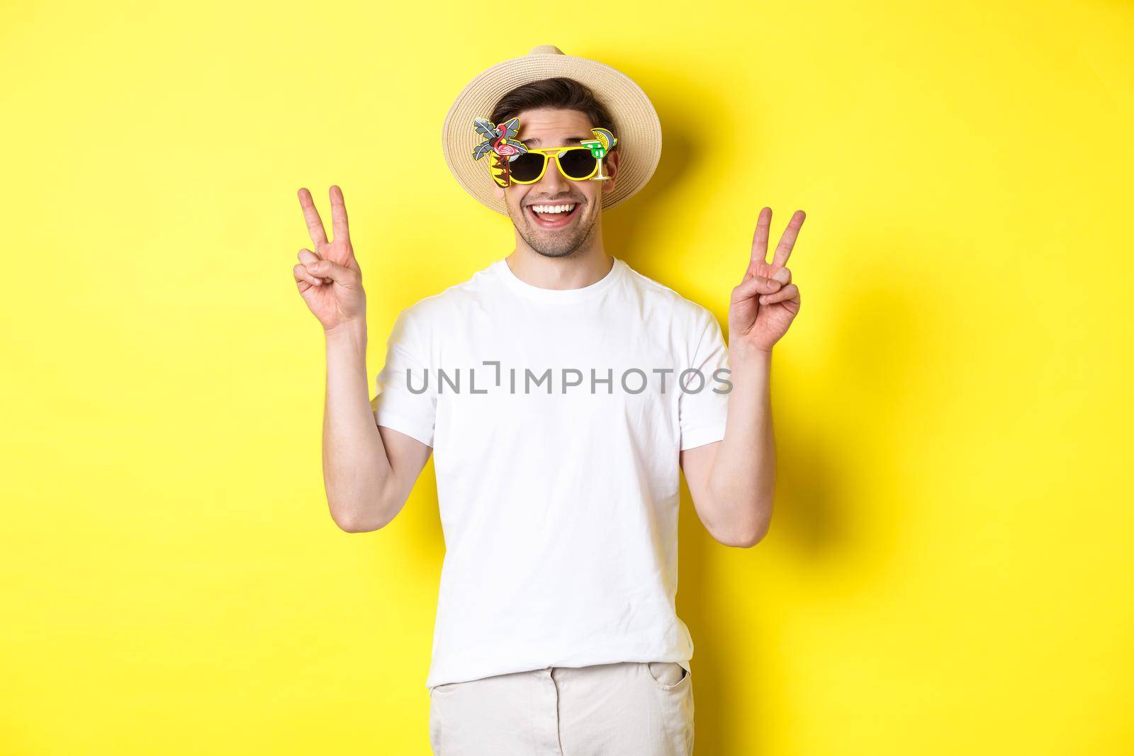 Concept of tourism and lifestyle. Happy man enjoying trip, wearing summer hat and sunglasses, posing with peace signs for photo, yellow background by Benzoix