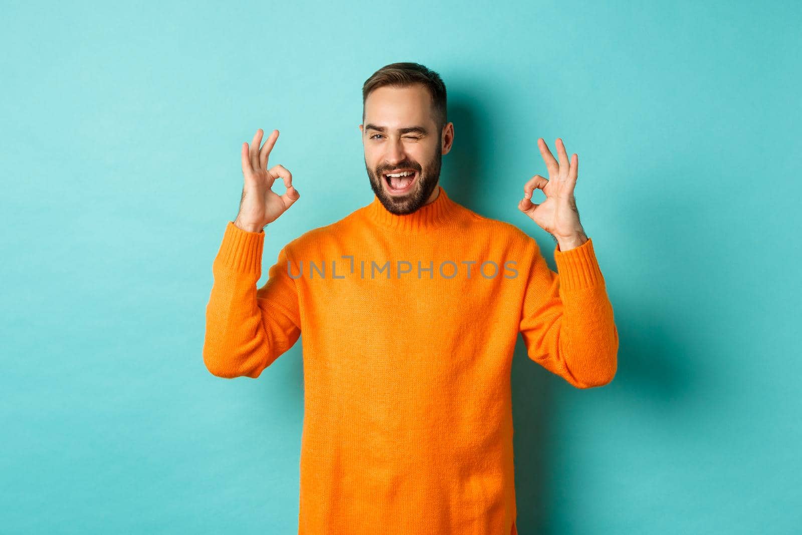 Confident man assuring all ok, showing okay signs and winking at you, recommending product, standing in orange sweater at studio background.