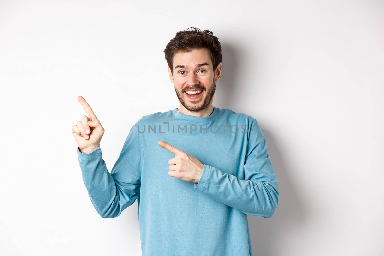 Happy caucasian man in casual blue sweatshirt, pointing fingers at upper left corner, showing link or logo on white background, smiling at camera by Benzoix