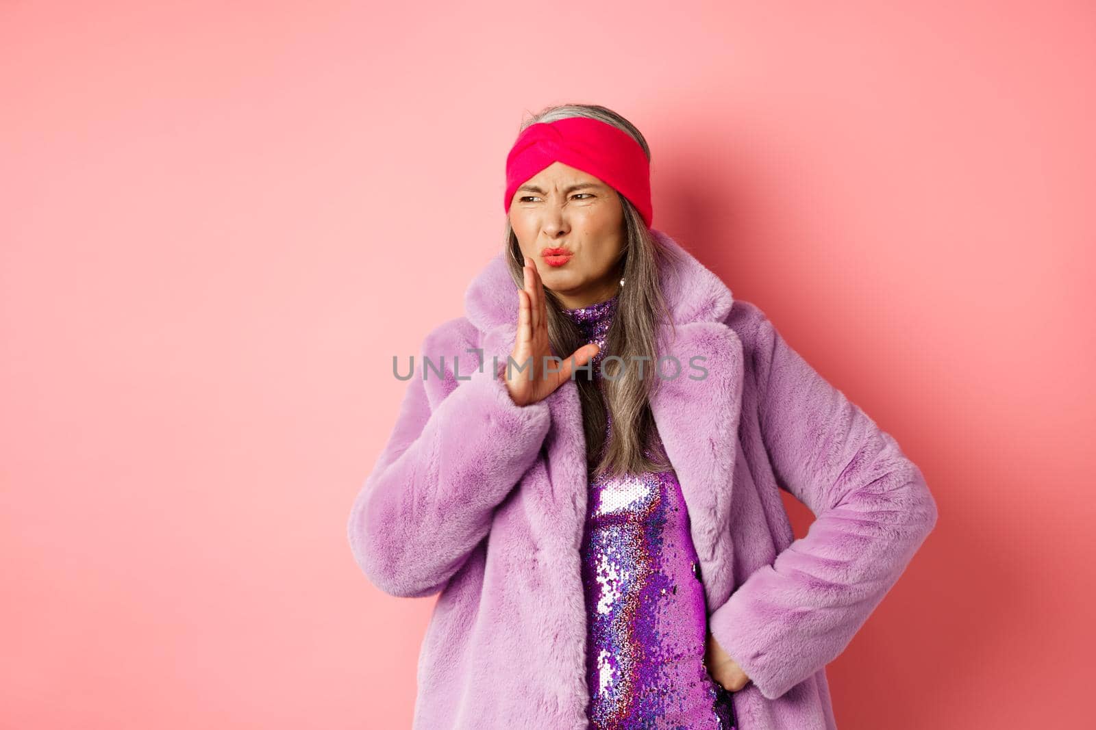 Fashion and shopping concept. Stylish asian middle-aged woman in fashionable winter clothes smell something bad, waving hand near nose from disgusting stink, pink background by Benzoix
