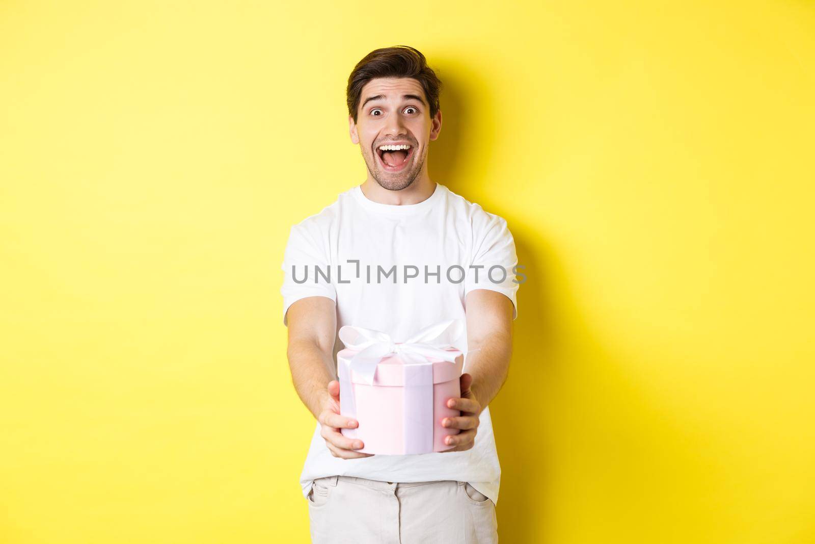 Concept of holidays and celebration. Happy man giving a gift and looking excited, standing against yellow background by Benzoix