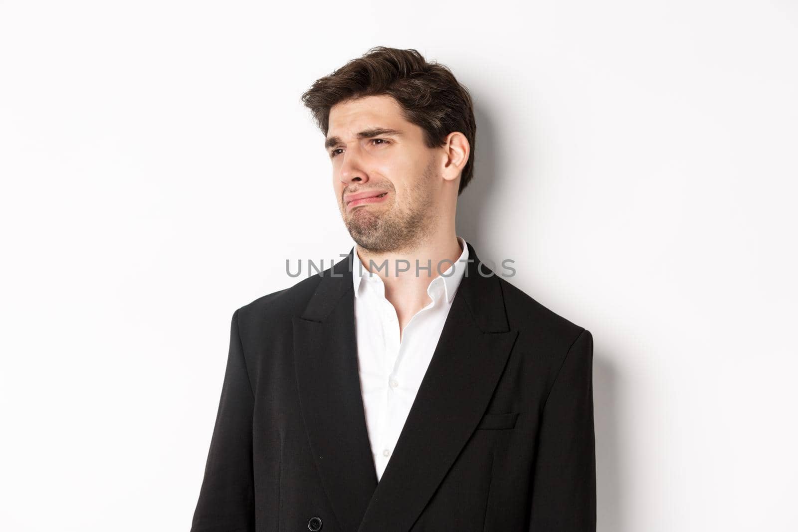 Close-up of disgusted young man in trendy suit, grimacing upset, looking left and standing against white background by Benzoix