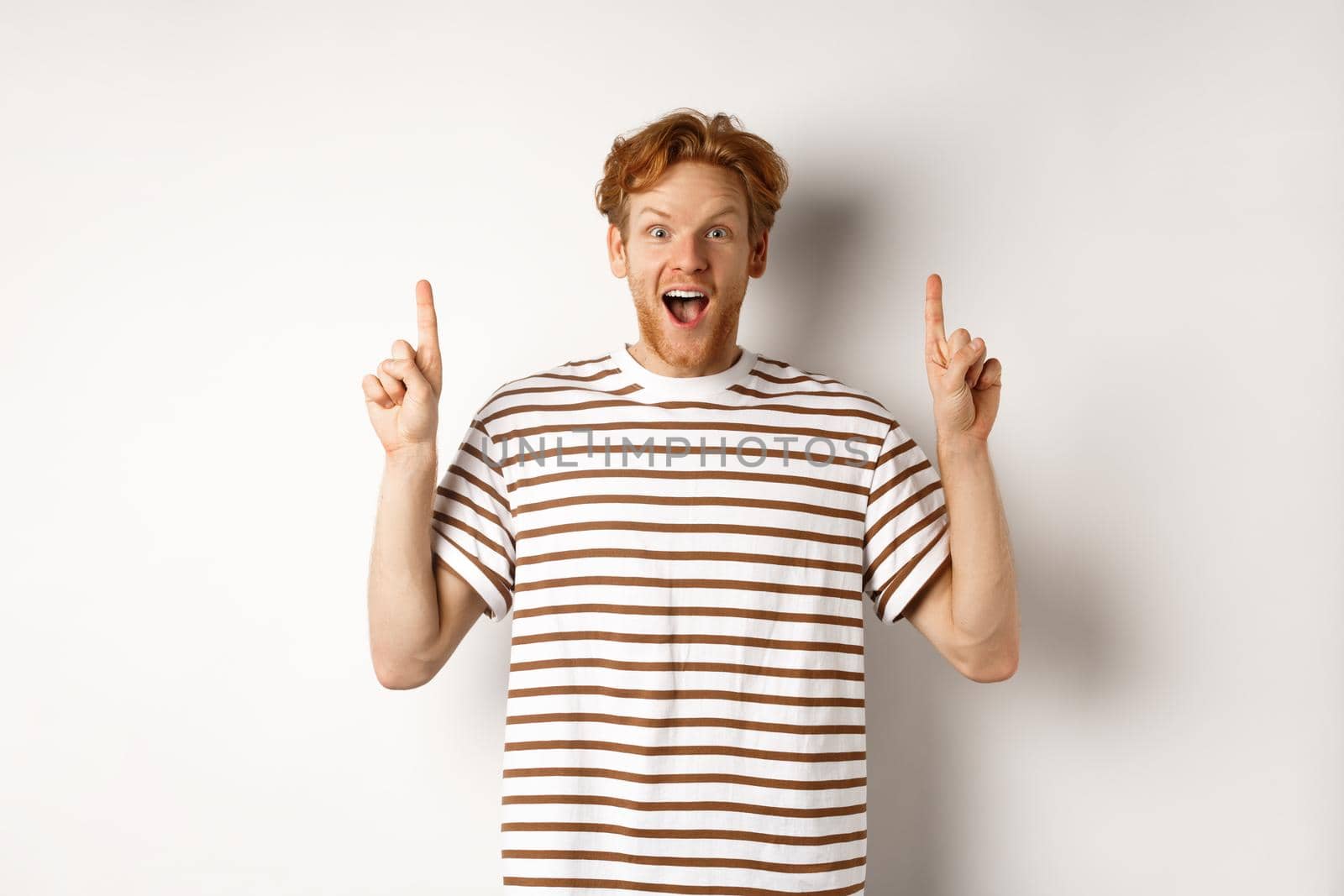 Amazed and excited redhead man checking out promotion, pointing fingers up and showing logo, staring at camera, standing over white background.