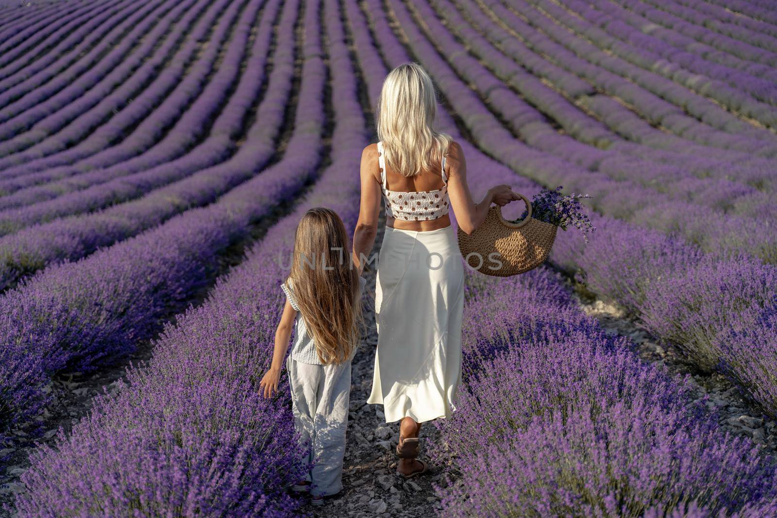 a beautiful girl with brown hair and a blonde girl in white dresses walk together through a lavender field.