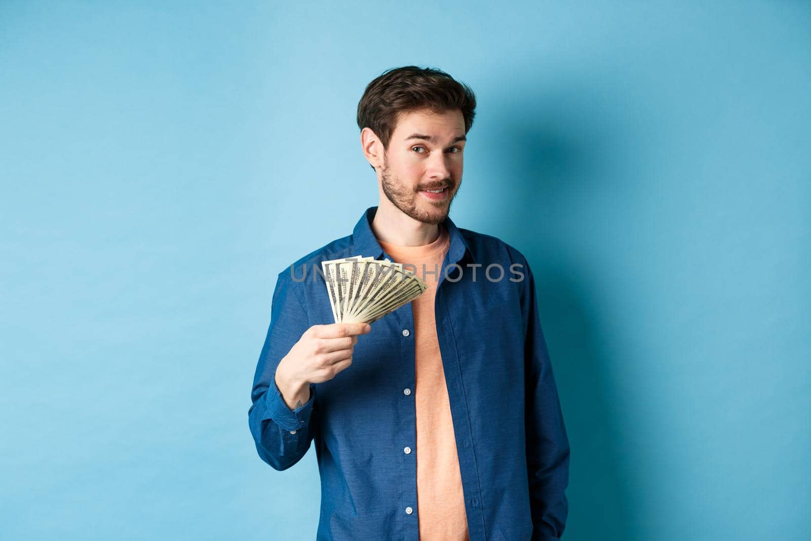 Unbothered rich guy showing cash and smiling, holding dollars in hand, standing on blue background by Benzoix