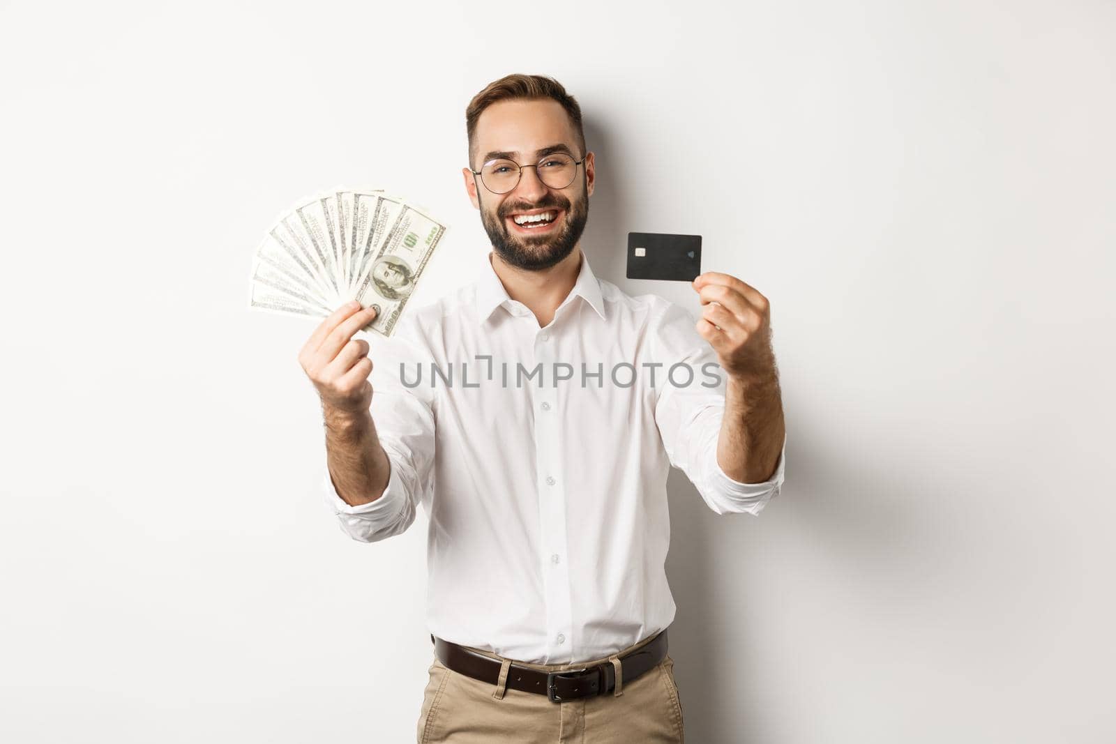 Happy young man showing his credit card and money dollars, smiling satisfied, standing over white background.