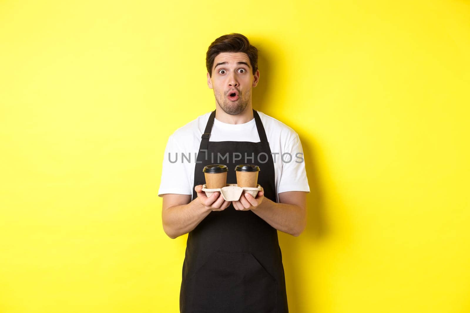 Barista serving coffee, looking surprised at camera, wearing black apron, standing against yellow background.