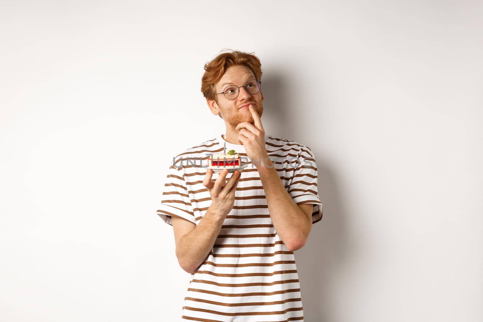 Holidays and celebration concept. Happy young man with red hair and glasses having birthday, holding cake with candle and thinking of b-day wish, standing over white background.