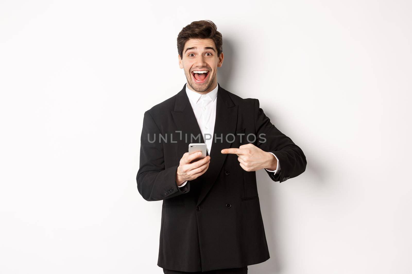 Image of cheerful businessman looking amazed, pointing at mobile phone, standing in suit over white background by Benzoix