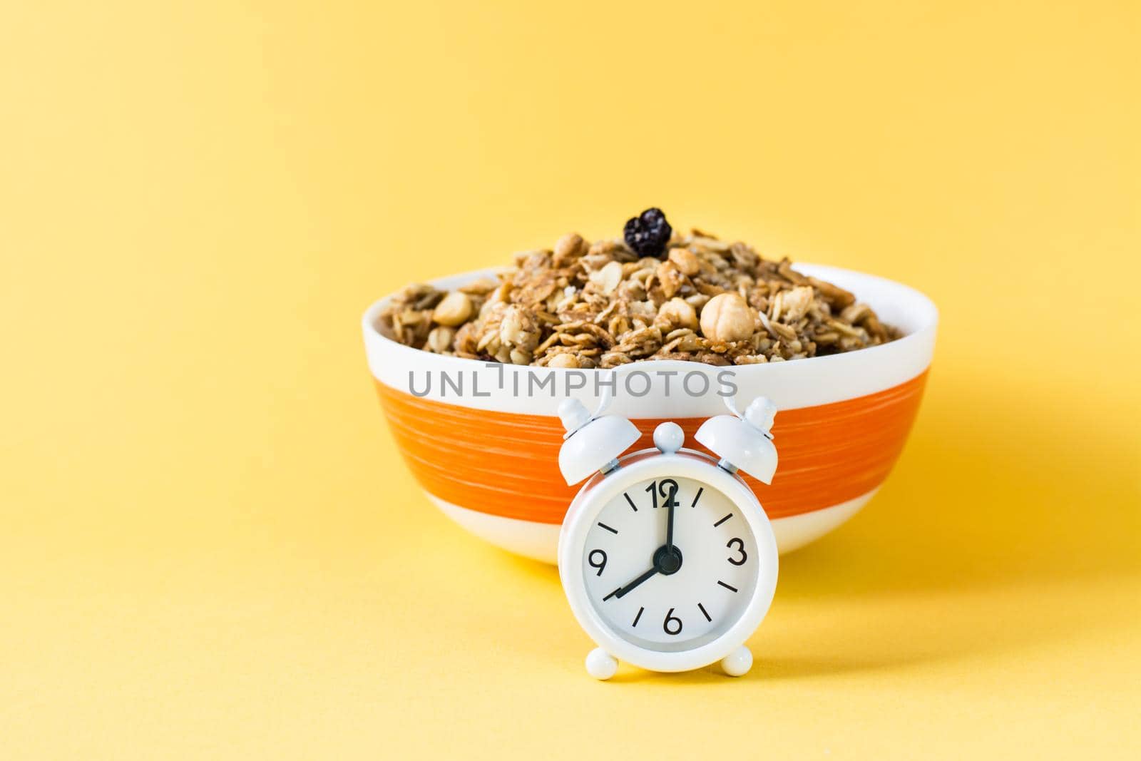 Healthy eating. Alarm clock in front of baked granola made from oats, nuts and raisins in a bowl on a yellow background by Aleruana