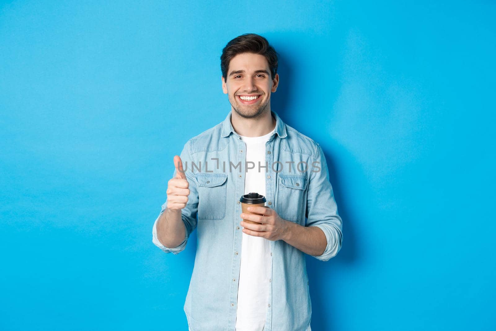 Handsome young man showing thumb-up and drinking coffee, recommending cafe takeaway, standing over blue background.