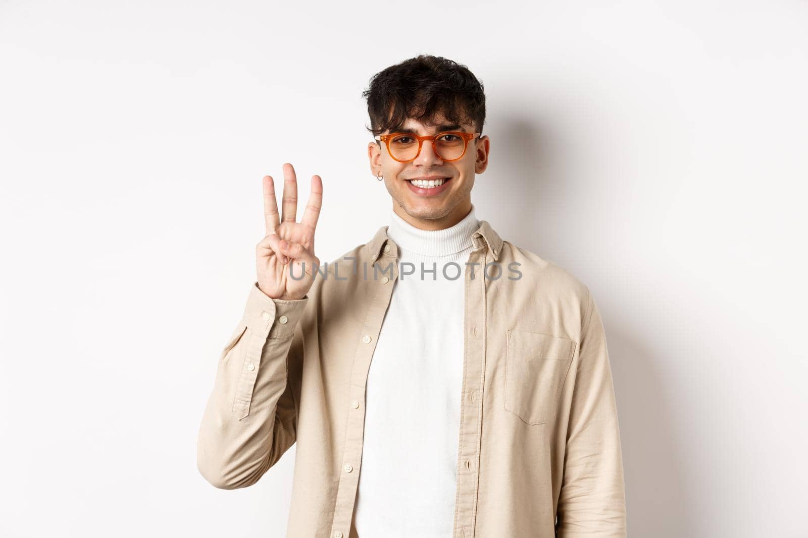 Handsome caucasian guy in glasses showing three fingers and smiling, standing on white background.