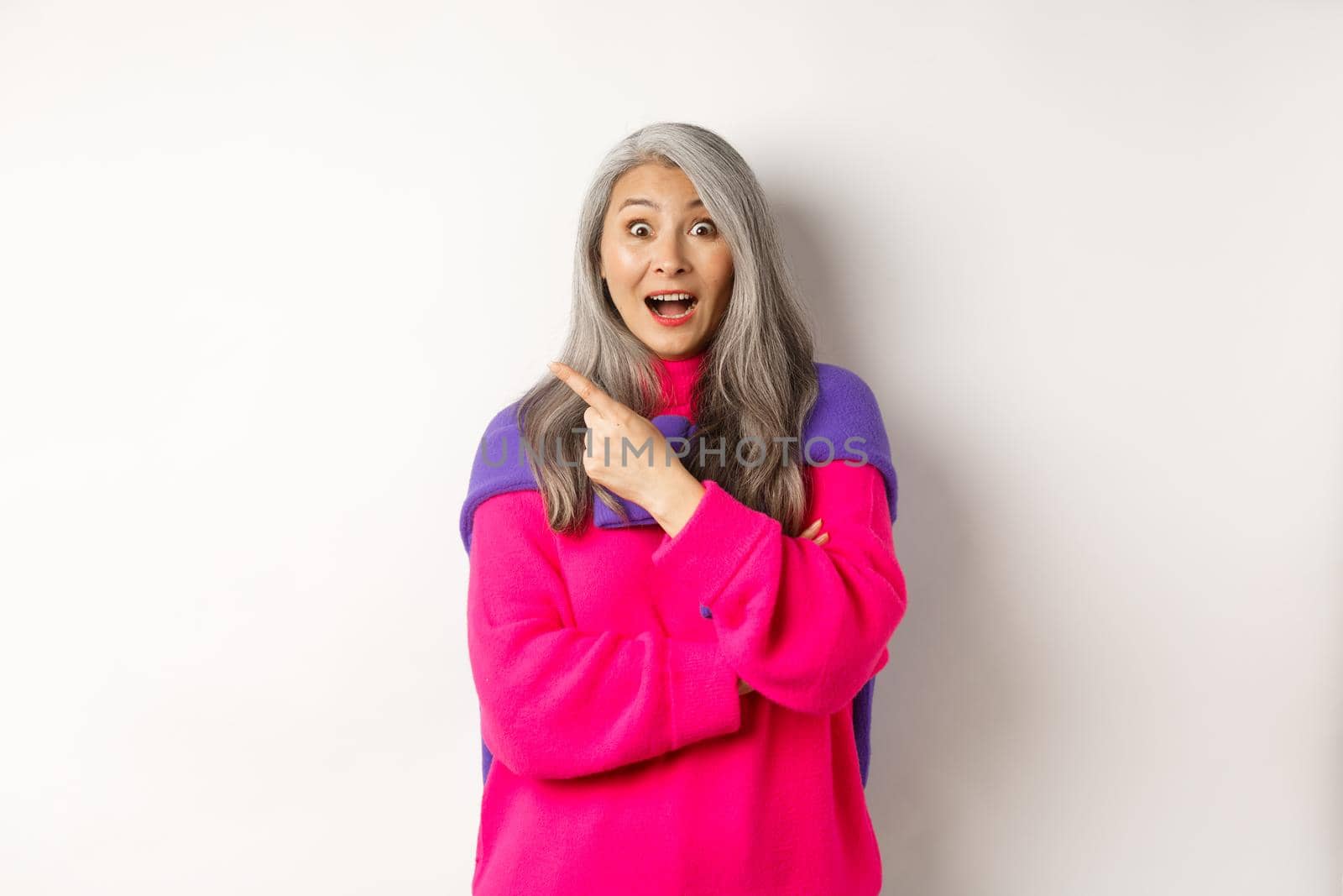 Portrait of impressed asian lady checking out promo offer, pointing at upper left corner and staring at camera amazed, standing in trendy pink sweater over white background.