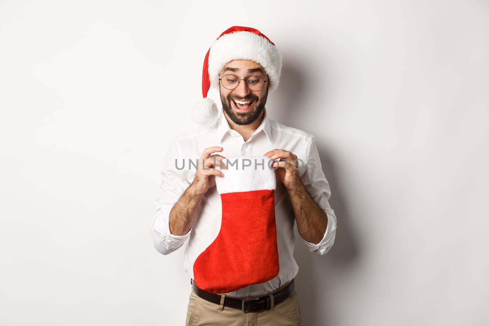 Happy adult man open christmas sock and looking inside, receiving xmas gifts for winter holidays, standing in santa hat, white background.