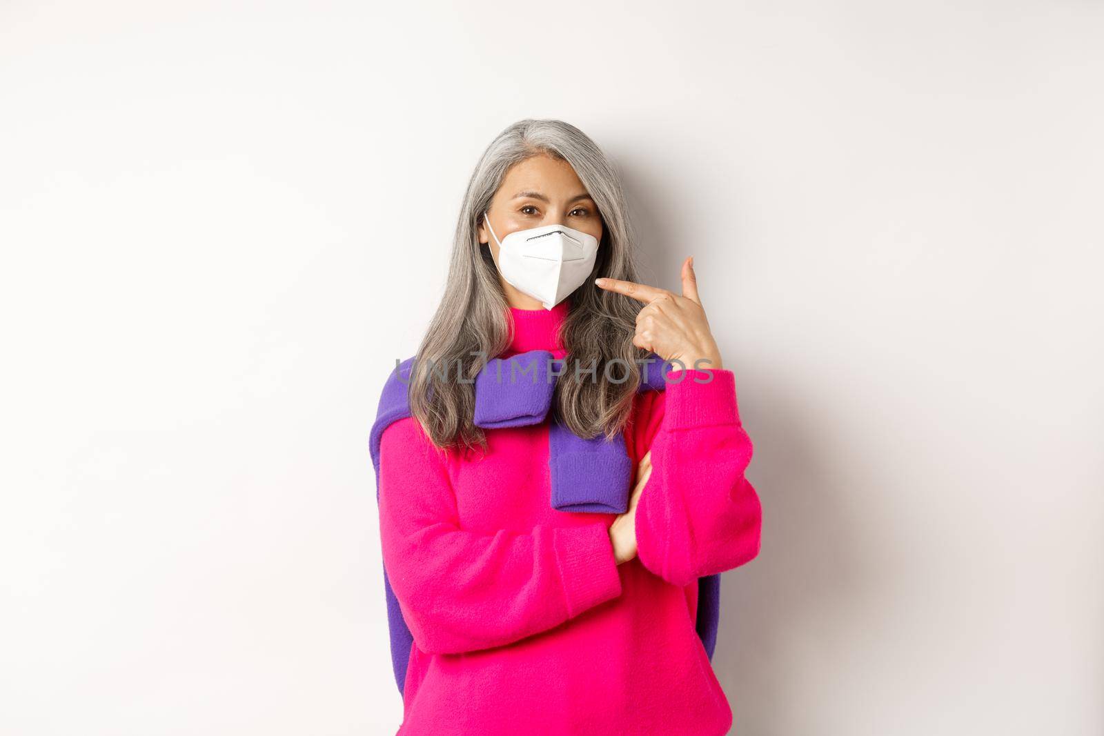 Covid, pandemic and social distancing concept. Fashionable asian senior woman wearing respirator, pointing at face mask and smiling, standing over white background by Benzoix