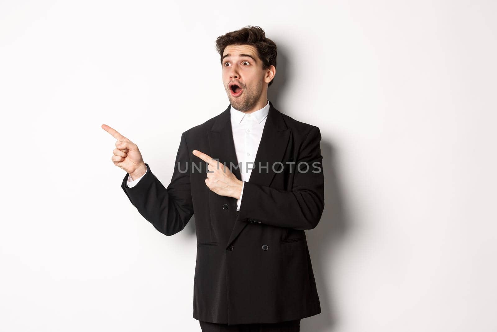 Impressed handsome man in party suit, looking at new year promo offer and pointing fingers left at banner, standing over white background by Benzoix